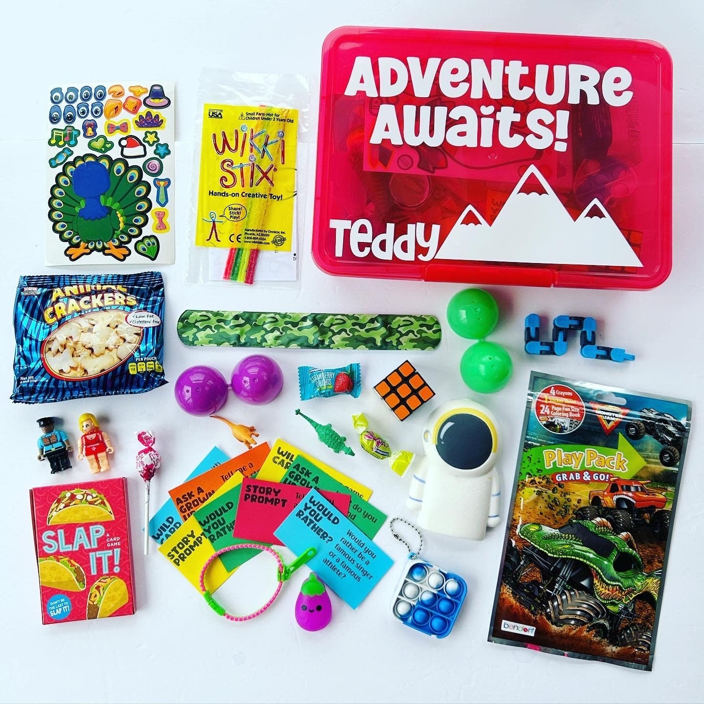 Red plastic box with adventure awaits wording, name Teddy, mountains. Around box are Wikki Stix, monster truck coloring set, activity cards, Slap It card game, fidgets, stickers, snacks, easter egg, lego mini figures, 2 dinosaurs, astronaut squishy