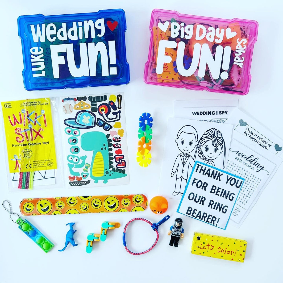 Two blue and pink plastic containers with the words 'Wedding Fun!' and 'Big Day Fun!' on them. The containers are filled with various small toys and activities, including a dinosaur sticker sheet, a wedding I Spy game, a word search, a ring bearer thank you card, a pop-it toy, a dinosaur toy, a Wikki Stix craft kit, and a coloring book.