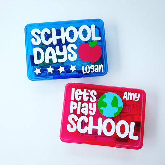 Two plastic containers with colorful designs and text that says "school days" and "let's play school" personalized with the names Logan and Amy. The blue container has the text 'SCHOOL DAYS' with a red apple and four stars. The red container has the text 'Let's Play SCHOOL' with a blue globe. Both containers have a name at the bottom, 'Logan' and 'Amy'. Filled with items for children to play pretend school making this a great gift for k