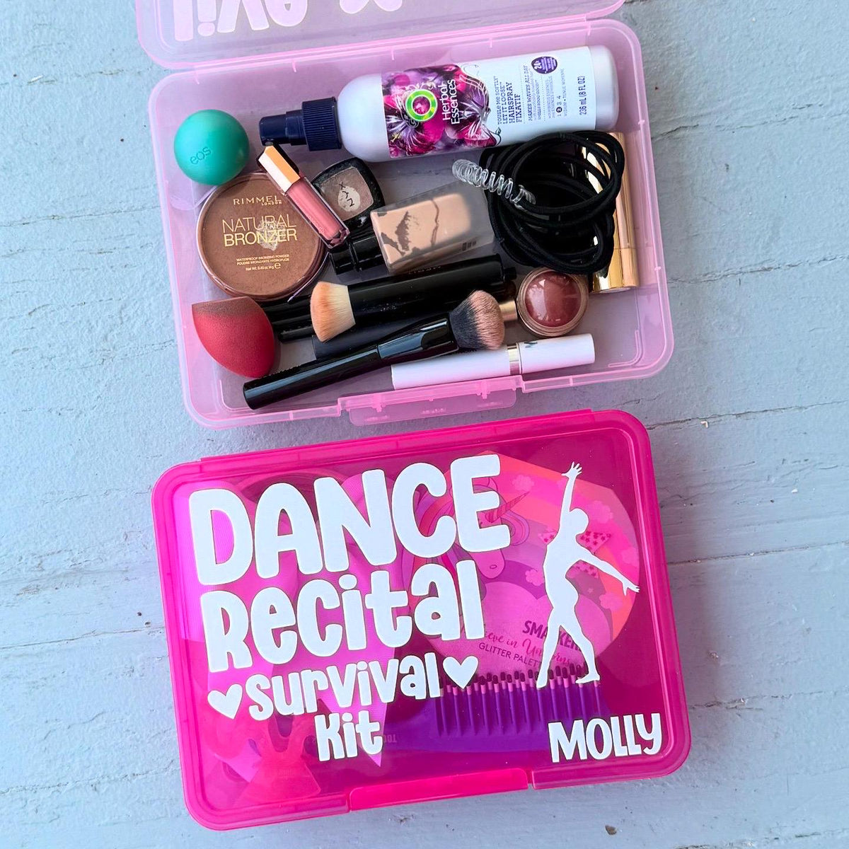 May include: Two pink plastic storage containers. The top container is open and filled with various items, including makeup brushes, hair ties, a hairbrush, a bottle of hairspray, and a compact. The bottom container is closed and has the words 'Dance Recital Survival Kit' and 'Molly' written on it in white letters. The container also has a silhouette of a dancer on it.