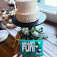 White wedding cake with decorative elements on a table, held by a person with a blue plastic box that says "wedding fun" and the name Teddy. Box is filled with children's activities to keep kids entertained at a wedding.