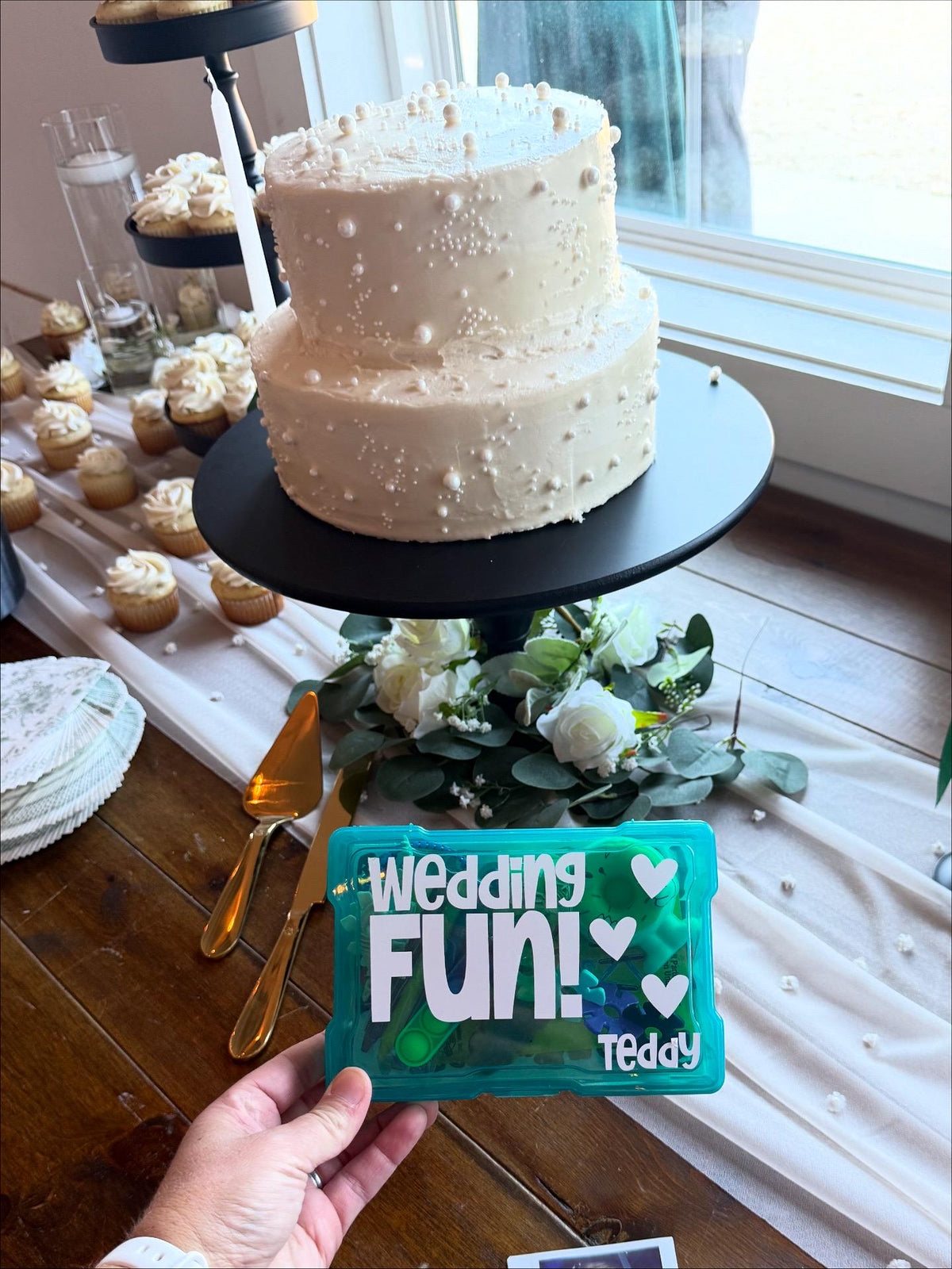 White wedding cake with decorative elements on a table, held by a person with a blue plastic box that says "wedding fun" and the name Teddy. Box is filled with children's activities to keep kids entertained at a wedding.