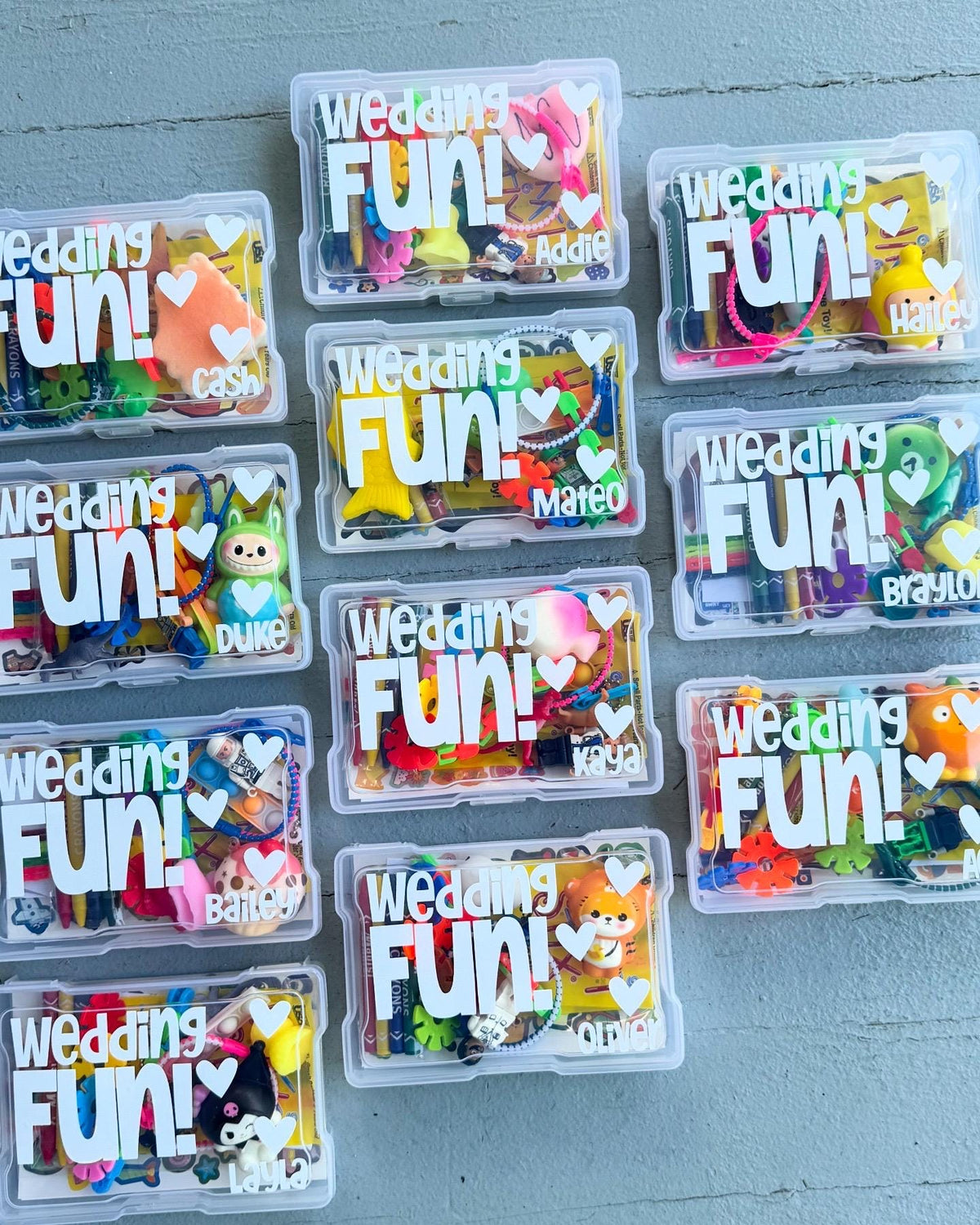 Small clear containers labeled 'Wedding Fun!' with colorful contents on a gray surface. Wedding activity kits for kids filled with wedding-themed activities for children, making these great wedding favors, flower girl or ring bearer gifts.
