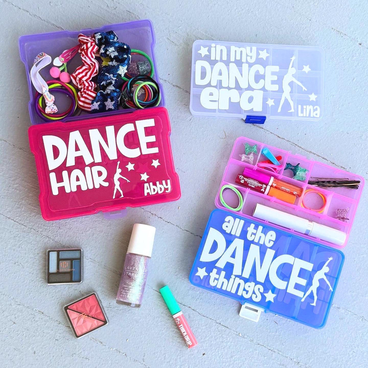 May include: Four clear plastic storage boxes with colorful lids. The boxes are labeled with dance-themed text and names. The boxes contain various items, including hair ties, makeup, and pens.