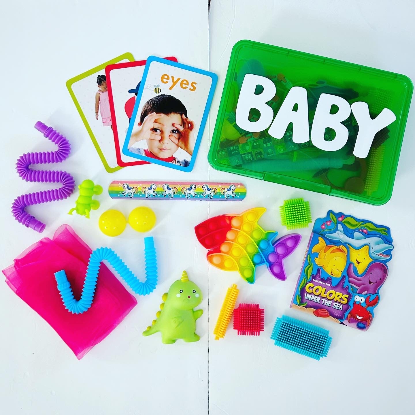 Green busy box with baby wording on it. Activities surround it including board book, pop it, pop tube, fidgets, suction cup toy, bristle blocks, scarf.