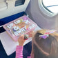 Young girl on an airplane with her airplane activity busy box on the airplane tray table. Light pink box with "up, up and away" and the name "Collins"