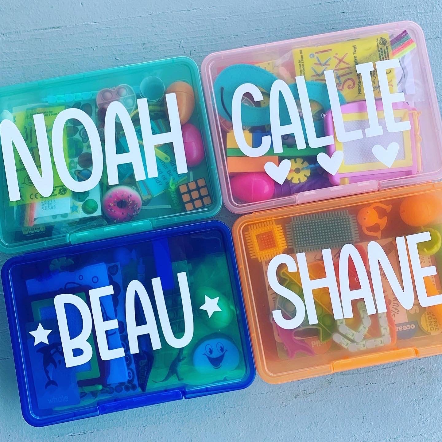 4 busy boxes for kids with personalization including turquoise with name Noah, pink box with Callie with 3 small hearts, blue box with Beau with 2 stars and orange box with Shane.