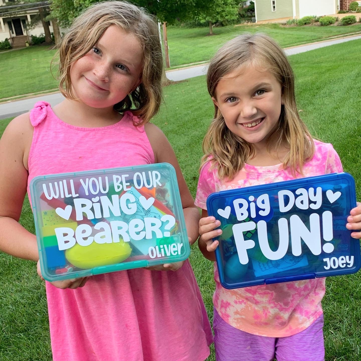 Two young girls holding wedding themed busy boxes. One girl holding a turquoise box with will you be our ring bearer question on the front. Other girl holding a dark blue box with big day fun and the name Joey on it.