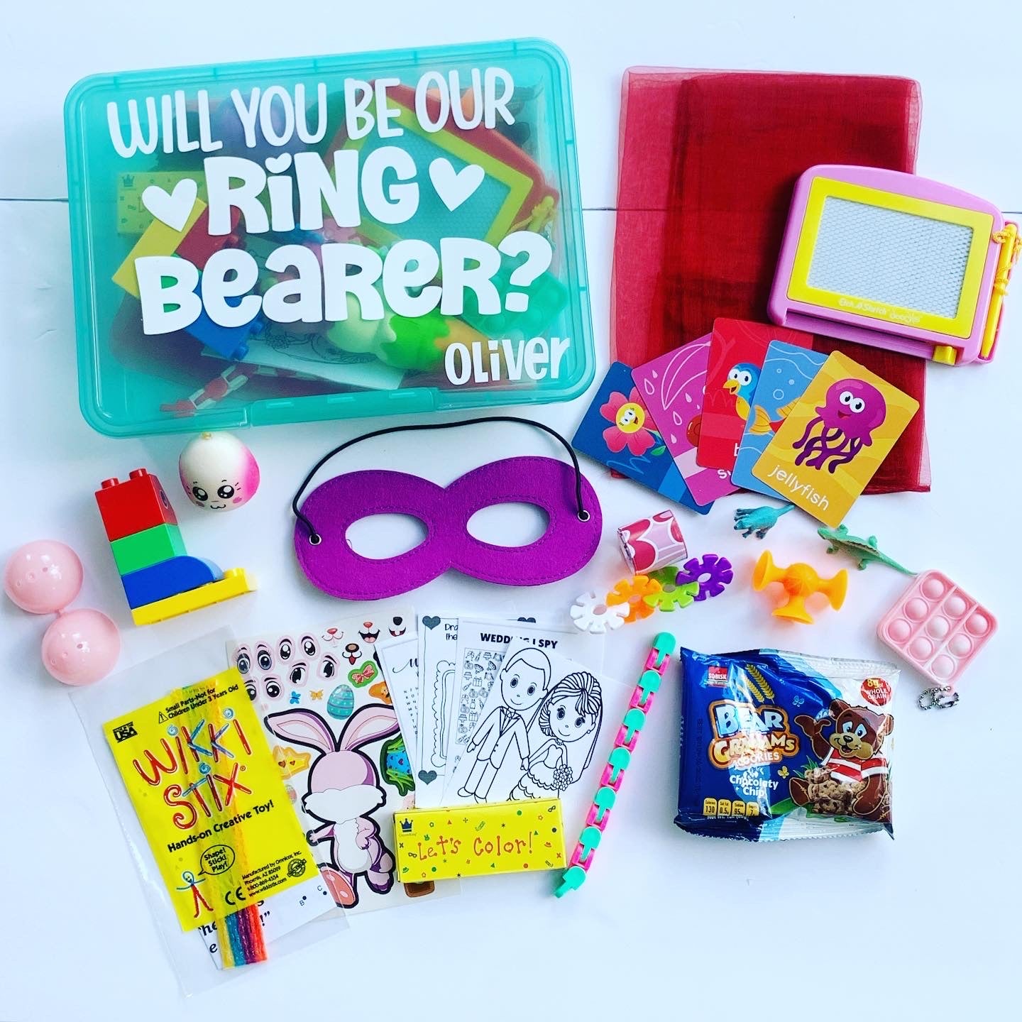 turquoise wedding busy box with will you be our ring bearer wording, name Oliver and two hearts. Activities surround box including peekaboo scarf, magna doodle, flashcards, slap bracelet, pop it keychain, teddy graham snacks, wedding coloring pages, crayons, wiki six, legos, squishy, dinosaurs, mask, fidgets